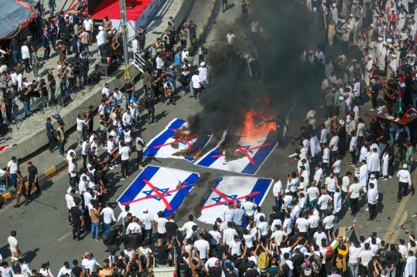 A large crowd in the street surrounds 4 Israel flags, two of which are on fire, as black smoke billows up from the flames.